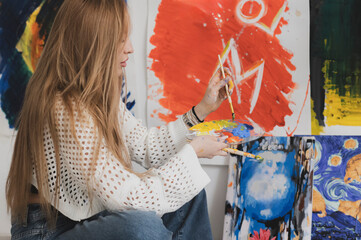 A female artist sits in a workshop, painting on canvas. She is wearing a white knit sweater and blue jeans. She is holding a paintbrush in her right hand and a palette in her left hand