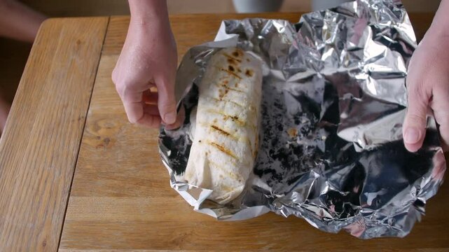 Hands unwrapping a grilled burrito from aluminum foil on a wooden table. The lightly charred tortilla reveals a freshly prepared, warm meal, food ready to be enjoyed