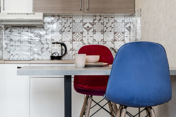 small simple kitchen in the apartment. White kitchen furniture, refrigerator, microwave, soft chairs in blue and red colors. Affordable real estate. There are plates on the table. Cozy dinner at home