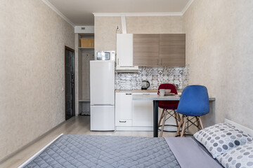 small simple apartment with kitchen and big bed . White kitchen furniture, refrigerator, microwave, soft chairs in blue and red colors. Affordable real estate. plates on table. Cozy dinner at home