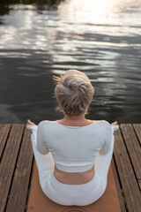 Fototapeta premium Mature woman in a white suit in a lotus position on the river bank