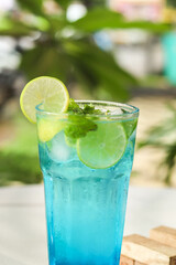 close up of Healthy Blue Lagoon Mojito soda served in glass with lemon and mint on a white table with jenga game and nature background. 