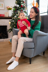 Mother is sitting in an armchair, holding her infant grandson, playfully chatting with him in the living room, which is decorated for the upcoming Christmas celebration