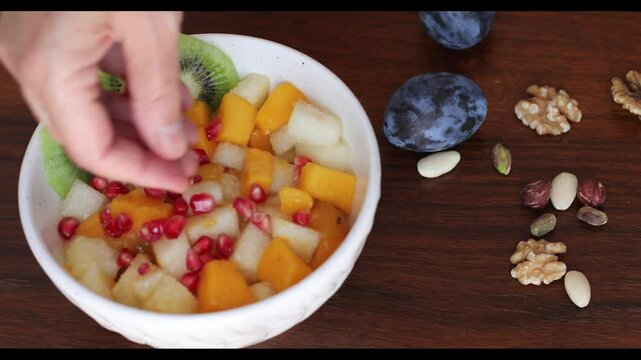Close-up of a ceramic bowl with fruit salad to which pomegranate seeds are added. On the side, dried fruit and plums.