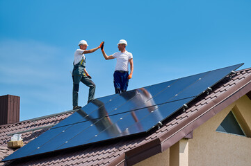 Worker building solar panel system on metal roof of house. Engineers giving high five to each...