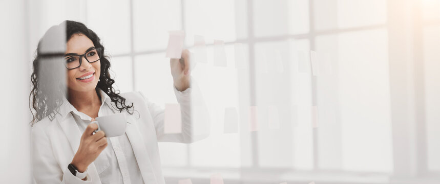 Business Concept. Smiling mexican businesswoman checking sticky notes glued to glass wall, drinking coffee at office