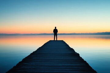 Silhouette of a person standing on a pier gazing at the calm water at sunrise