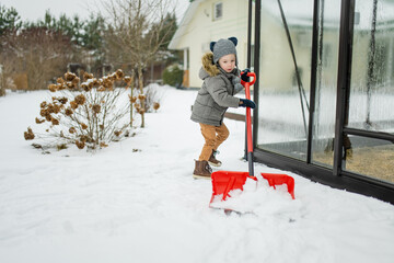 Adorable little boy helping to shovel snow in a backyard on winter day. Cute child wearing warm...