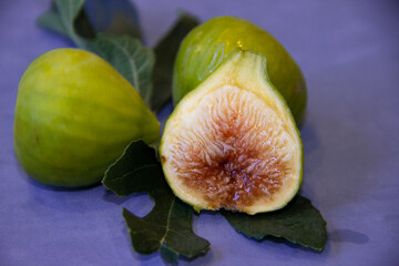 Ripe green figs accompanied by their leaves placed on a plush blue surface and a juicy cut fig with...