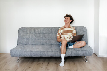 Young man sits casually on a gray sofa, smiling while using a laptop in a bright indoor living space