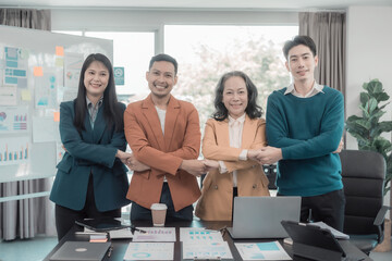 Asian businessman stands with his arms crossed across his chest and smiles confidently, while successful colleagues hold hands, showing unity in a conference room.