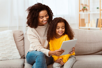 Cute Black Family Of Mother And Daughter Using Tablet Computer Watching Cartoons Together Sitting On Couch At Home.