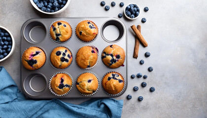 Delicious freshly baked blueberry muffins arranged in a muffin tray, surrounded by fresh blueberries and cinnamon sticks.
