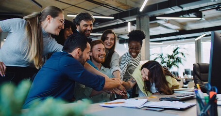 Multi-ethnic group of people gathering together in front of one computer. Everyone raising their hands together. Team building. Happy about positive outcome after discussion.