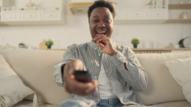 Portrait of cheerful African man watching TV and laughing, switching channels. Young black guy enjoying comedy and sitcom on television, leisure in weekend, spending time alone in apartment, pastime