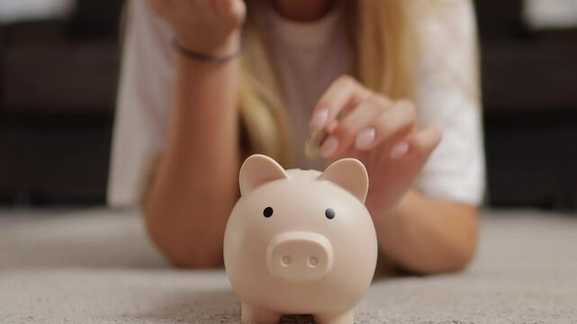 Young girl lies on carpet in her living room, putting coins into piggy bank. scene reflects focus on saving money and planning for future financial goals
