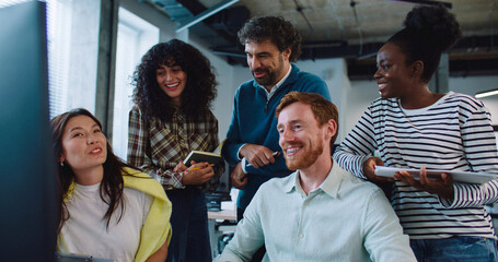 Positive multi-ethnic team working together at one desk. Listening to young man in middle describing something on screen and making gestures with his hand. Everyone looking at camera and smiling.