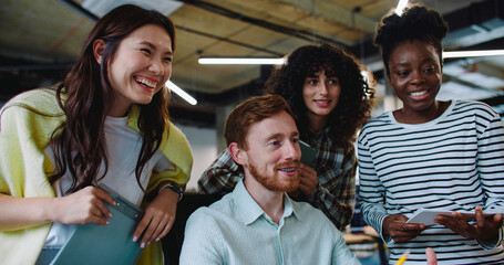 Handsome Caucasian man with beard and red hair sitting in front of big monitor. Three girls behind listening carefully to experienced male. Explaining something to newcomers. Pointing at screen.