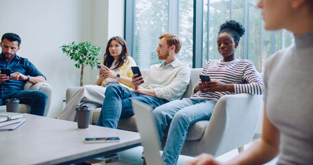 Obraz premium Multi-ethnic group of people sitting on soft couches. Everyone using gadgets. Woman in blurred vision close to camera typing on laptop keyboard. Coffee cup on table.