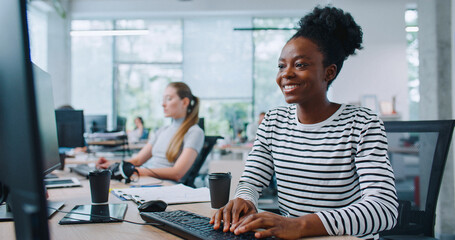 Arican American woman with plastic cup of coffee sitting down on office chair. Woman with smile starting to type message on computer. Working in agency or financial company.