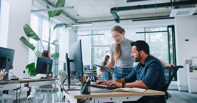 Beautiful Caucasian woman with clipboard standing next to worker in office. Employees discussing about work issues. Busy man typing text on computer and intently listening to manager. Teamwork.