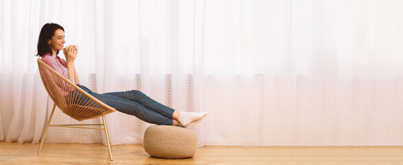 Morning Relaxation. Girl sitting in armchair putting legs on ottoman and enjoying coffee against...