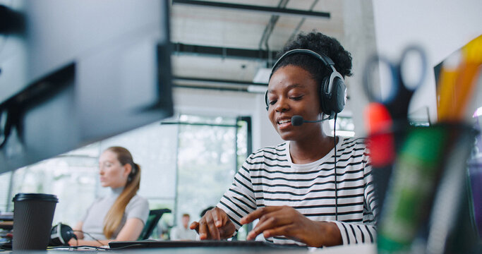 Camera view of diverse women sitting in front of computer displays. Worker on right side and wearing headset for remote consultation with clients. Working in call center. Customer support.