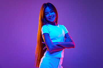 Casually dressed young girl, confidently crossed hands and looking at camera in neon light against gradient purple studio background. Concept of back to school, childhood, fashion and style. Ad