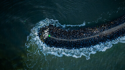 View from above of rocky pier extending into the ocean, marked by a green buoy with waves gently crashing against its sides. Aerial view of stone breakwater with green marker at entrance of harbor.