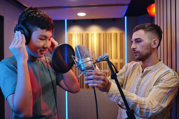 Two people engaging in recording session with microphone and headphones surrounded by professional studio equipment illuminated by ambient lights