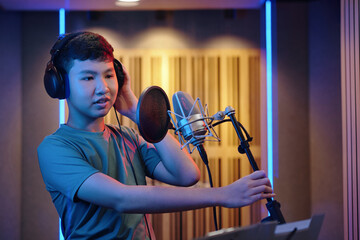 Young Asian boy in studio adjusting microphone wearing headphones and singing into pop filter with acoustic panels in background