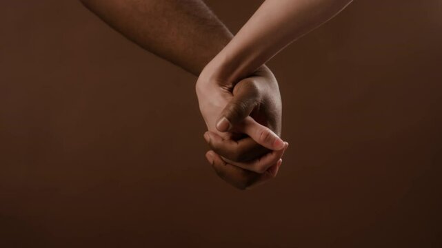 Close-up view of multi-ethnic couple holding hands with interlocked fingers showing unity on brown background