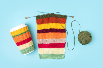 Cup for takeaway coffee with knitted autumn striped cover and knitted wool with knitting needles on turquoise background. Concept of takeaway coffee, walking, energetic life, hobbies needlework
