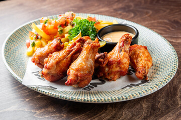 plate of crispy fried chicken wings, golden French fries, pickles, and a side of salsa, accompanied by a cup of dipping sauce, all served on a rustic wooden table. Perfect for a hearty meal or snack.