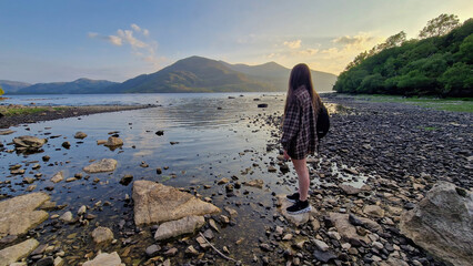 A woman stands at the edge of Muckross Lake, soaking in the breathtaking sunset that paints the sky...
