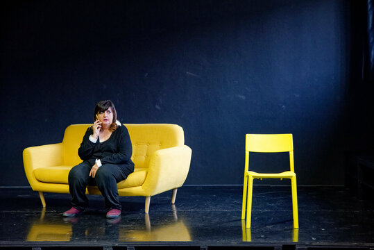 Woman sitting thoughtfully on a yellow sofa with an empty yellow chair beside her on a dark stage.