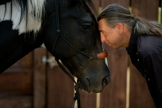 Man affectionately touches noses with a black horse in a stable setting.