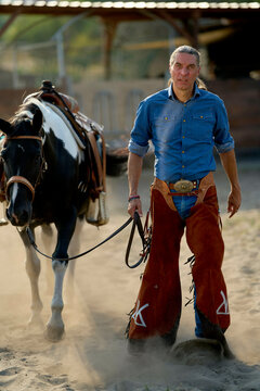 A confident man in cowboy attire walks beside a saddled horse in a dusty arena.
