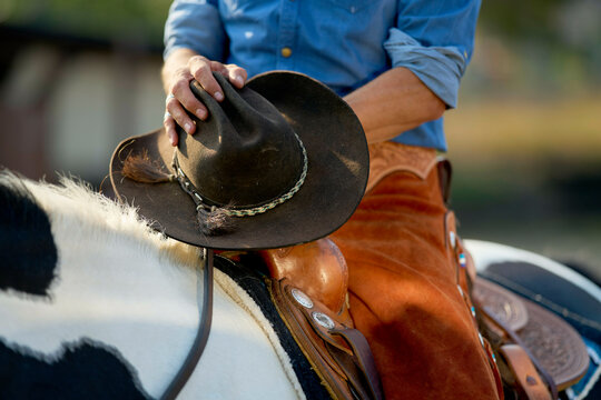 Cowboy in blue shirt adjusting a brown cowboy hat while riding a black and white horse.