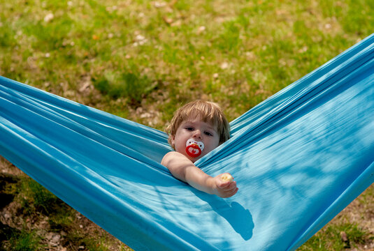 Toddler with a pacifier relaxes in a blue hammock, reaching out with one hand.