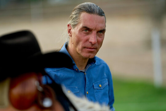 Man with grey long hair and blue shirt giving an intense stare behind a blurred figure in a cowboy hat.