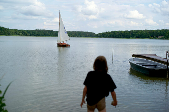 A child stands at the edge of a calm lake watching a sailboat glide by, with docked rowboats nearby.