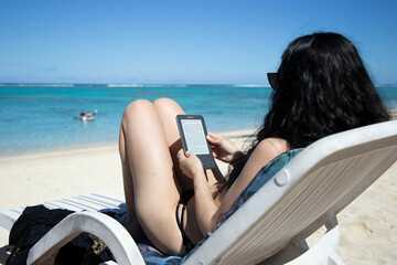 Woman relaxing on a beach chair reading an e-book with turquoise waters and swimmers in the background, Titikaveka, Rarotonga, Cook Islands