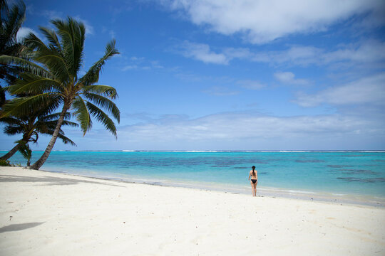 Woman in black Bikini walks along white sand beach towards crystal clear turquoise blue water on a tropical island beach, Titikaveka, Rarotonga, Cook Islands
