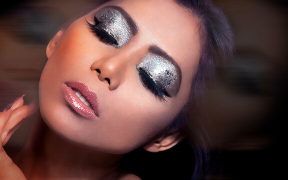 Close-up of a woman with dramatic glitter eyeshadow and glossy lips in soft-focus background.