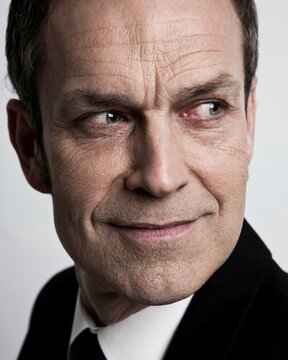 Close-up portrait of a smirking man in a black jacket against a white background.