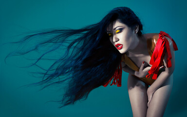 Woman with long flowing black hair and striking yellow eye makeup wearing a red harness on a teal background.