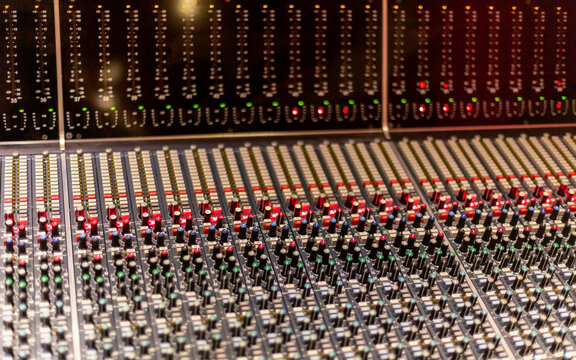 Close-up of a professional audio mixing console with numerous adjustment knobs and sliding faders illuminated by red and green indicator lights.