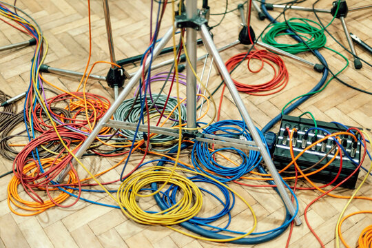 Colorful cables tangled on a wooden floor amidst music stands and microphone stands.