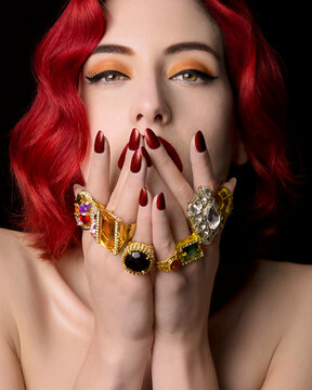 Red-haired woman with vibrant makeup showcasing an assortment of colorful, gemstone-encrusted rings and bracelets